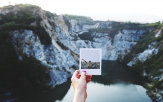POV shot of a an outstretched hand holding a polaroid photo while standing in the mountains