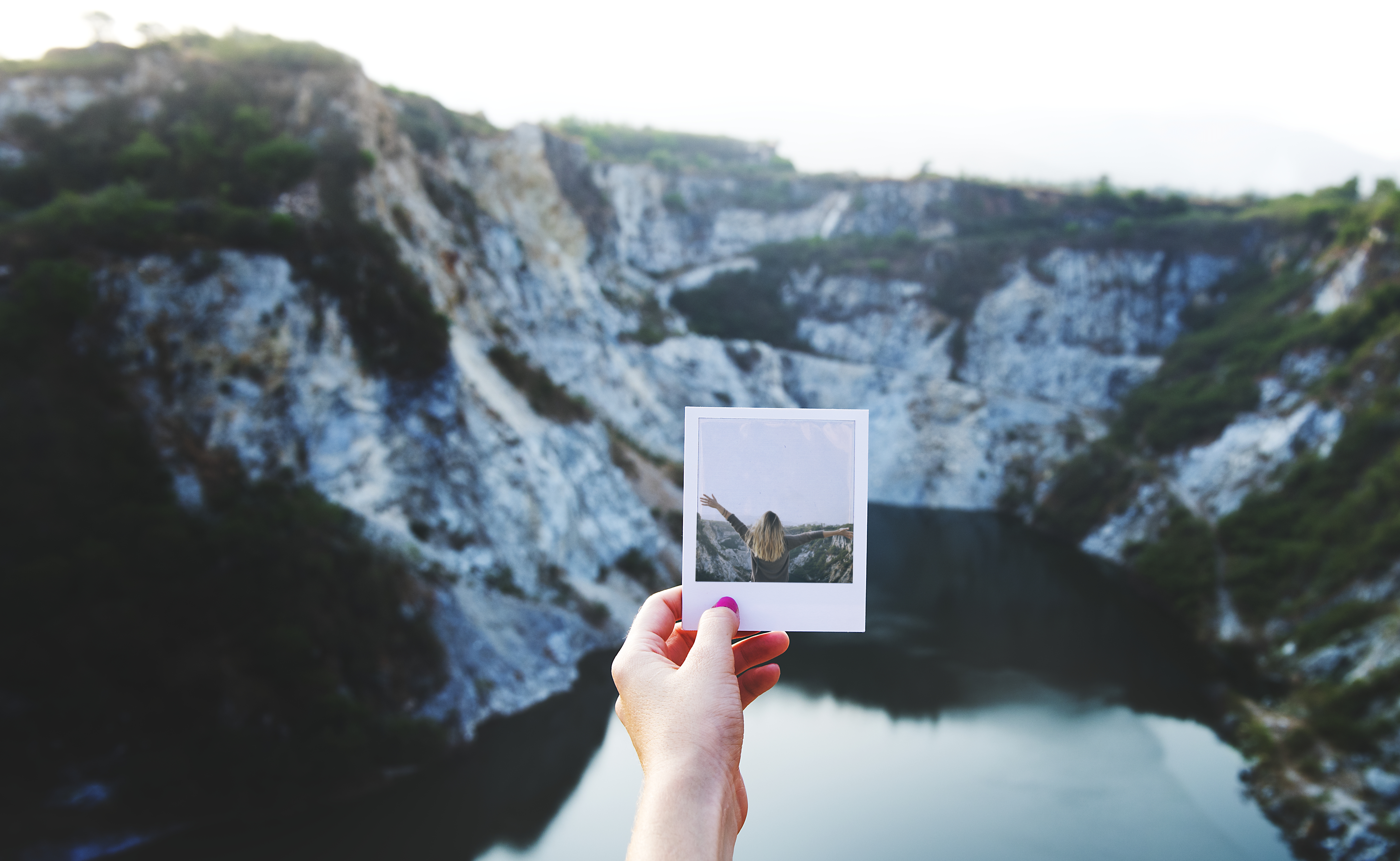 POV shot of a an outstretched hand holding a polaroid photo while standing in the mountains