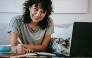 Girl with a pen in hand, looking at a computer with a cat beside her
